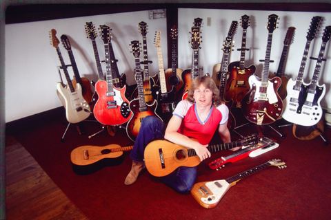 A musician sits among various guitars and instruments in a cozy room.