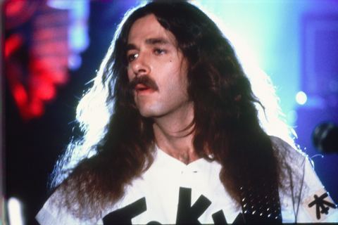 Musician with long hair and a mustache performs against a colorful, blurred background.