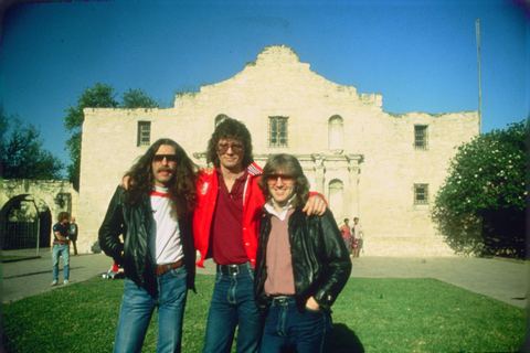 Three men pose together in front of a historic stone building under a clear blue sky.